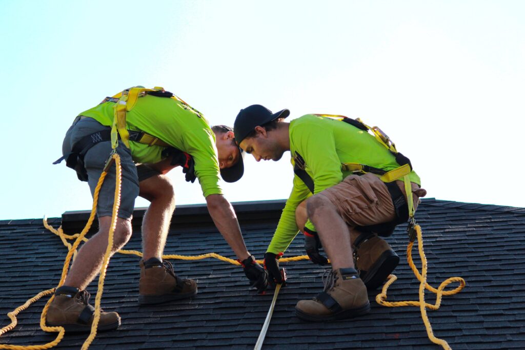 Two workers wearing safety gear install architectural shingles on a roof
